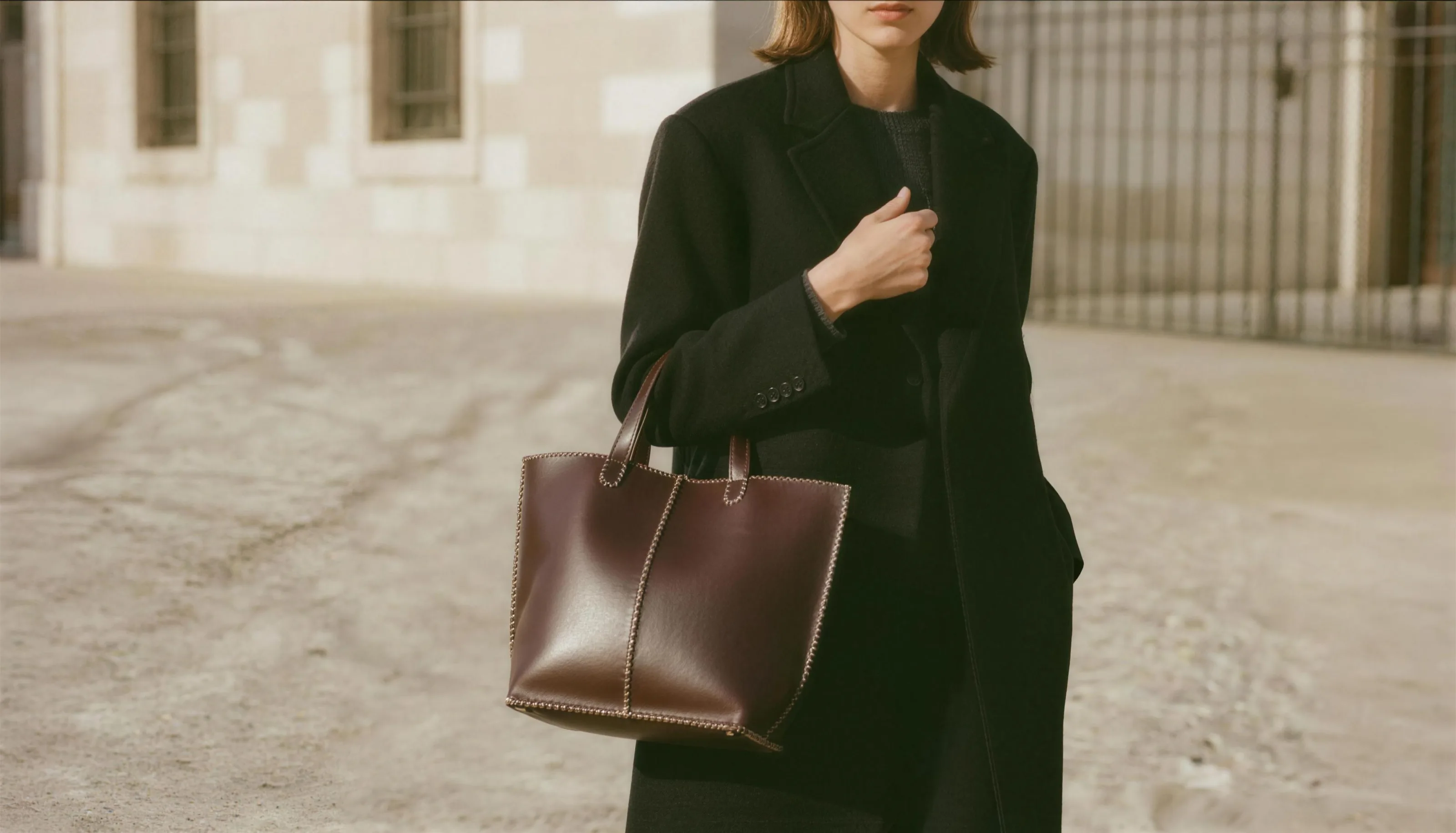 An oxblood leather tote with cream contrast stitching, carried against a black wool coat on a warm pale-stone street