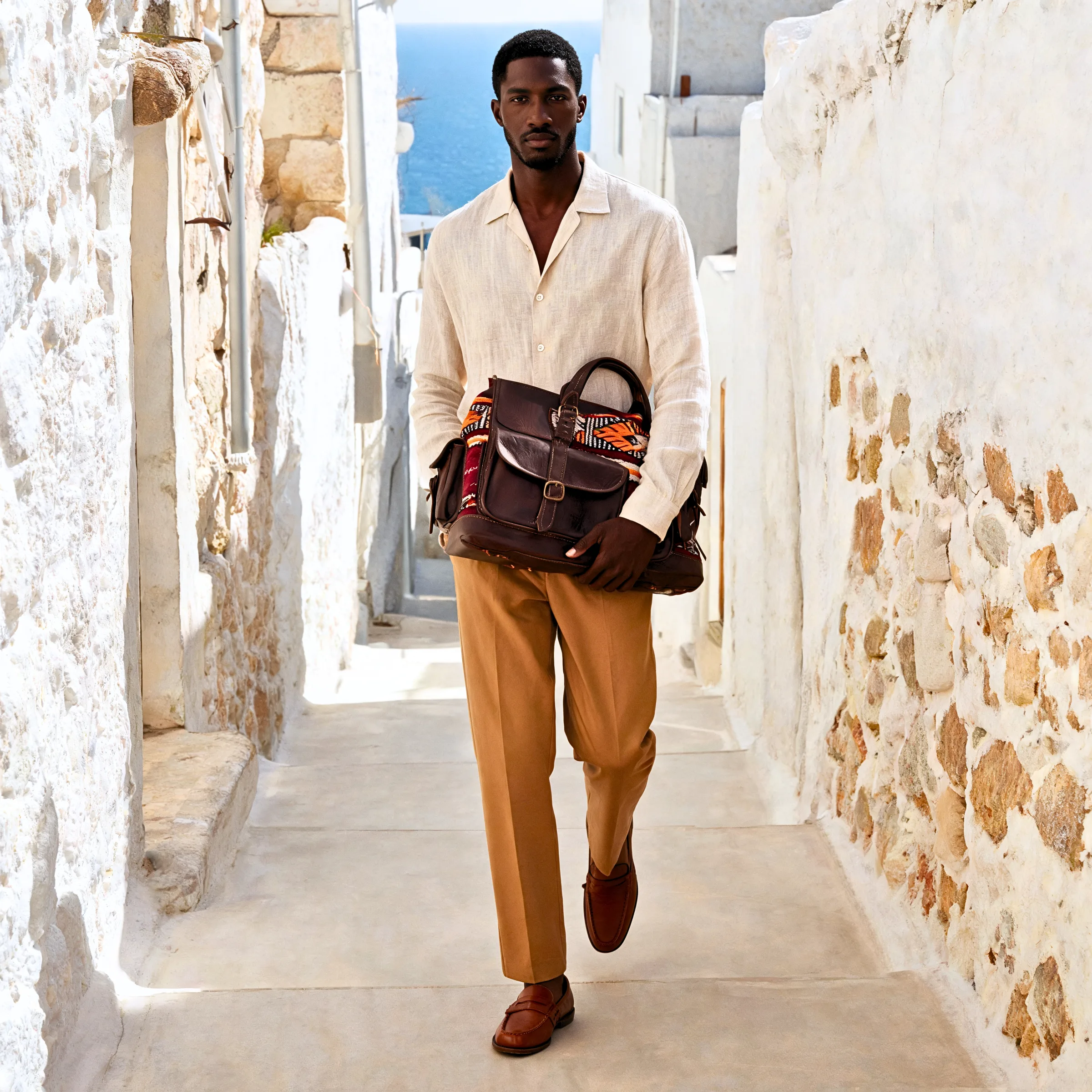 A man in cream linen carries a leather bag down a whitewashed Mediterranean lane, the sea beyond